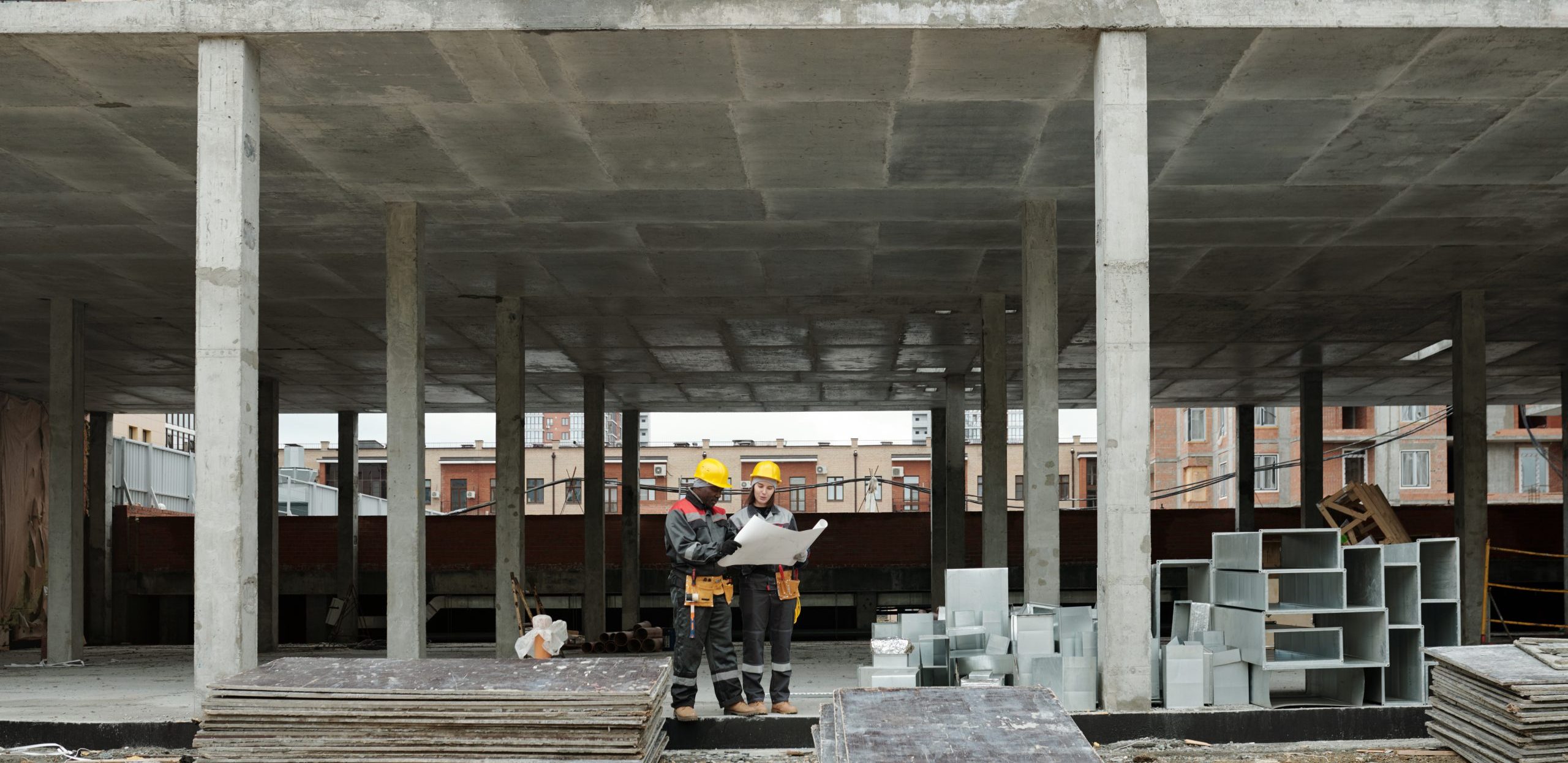 Two interracial builders in workwear discussing sketch of new building at construction site while standing inside unfinished concrete structure